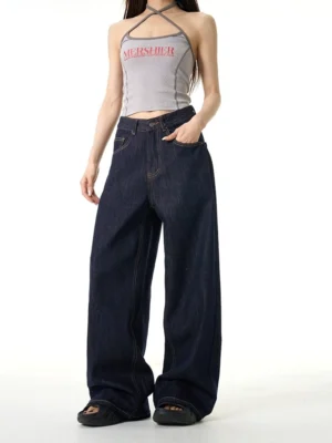 Female model wearing mid-rise dark wash wide-leg streetwear jeans with a gray printed halter top and black open-toe shoes in a minimalist studio