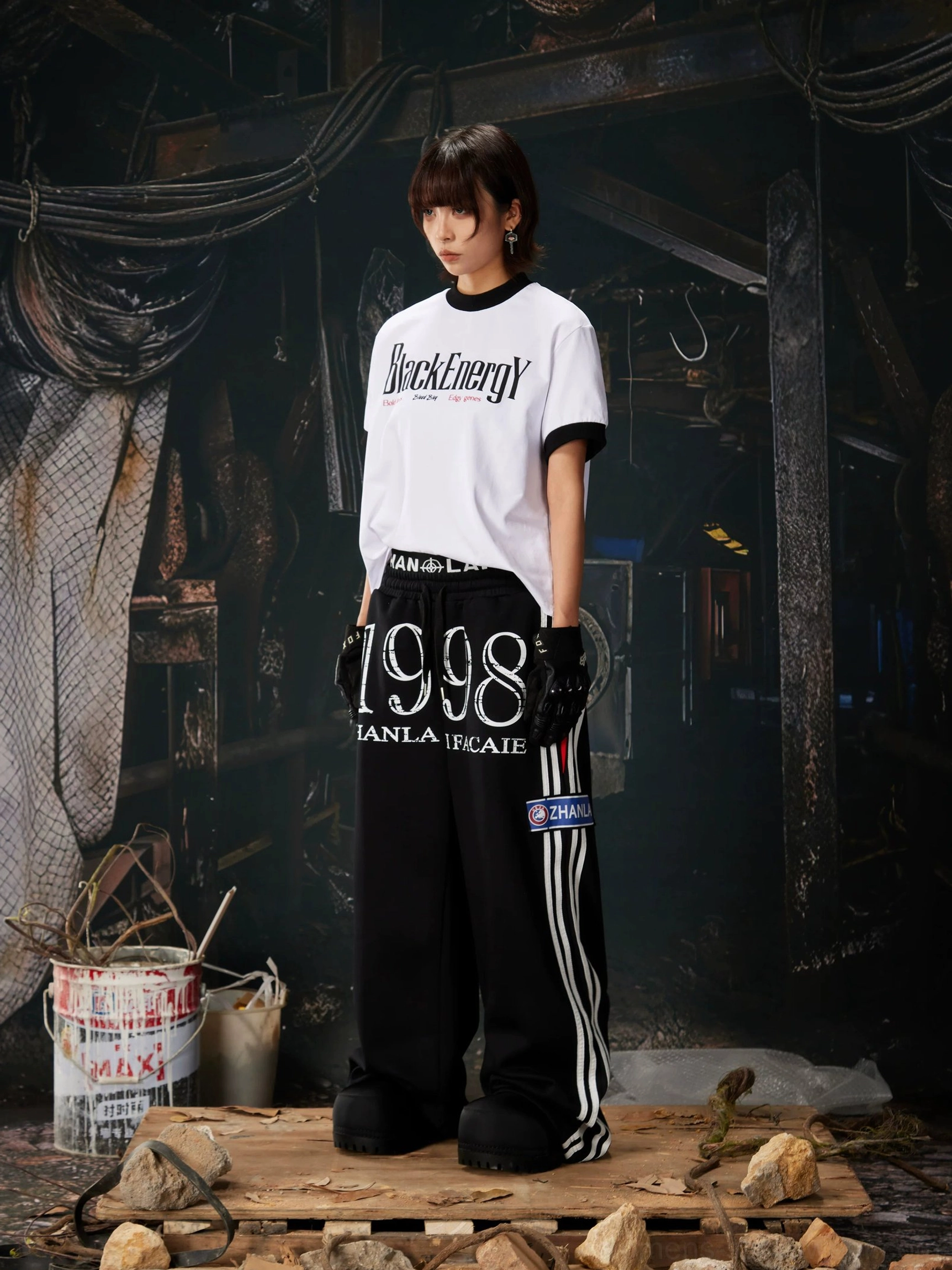 Black 1998 streetwear sweatpants styled with a cropped white graphic tee, wide-leg silhouette, and contrast athletic side stripes