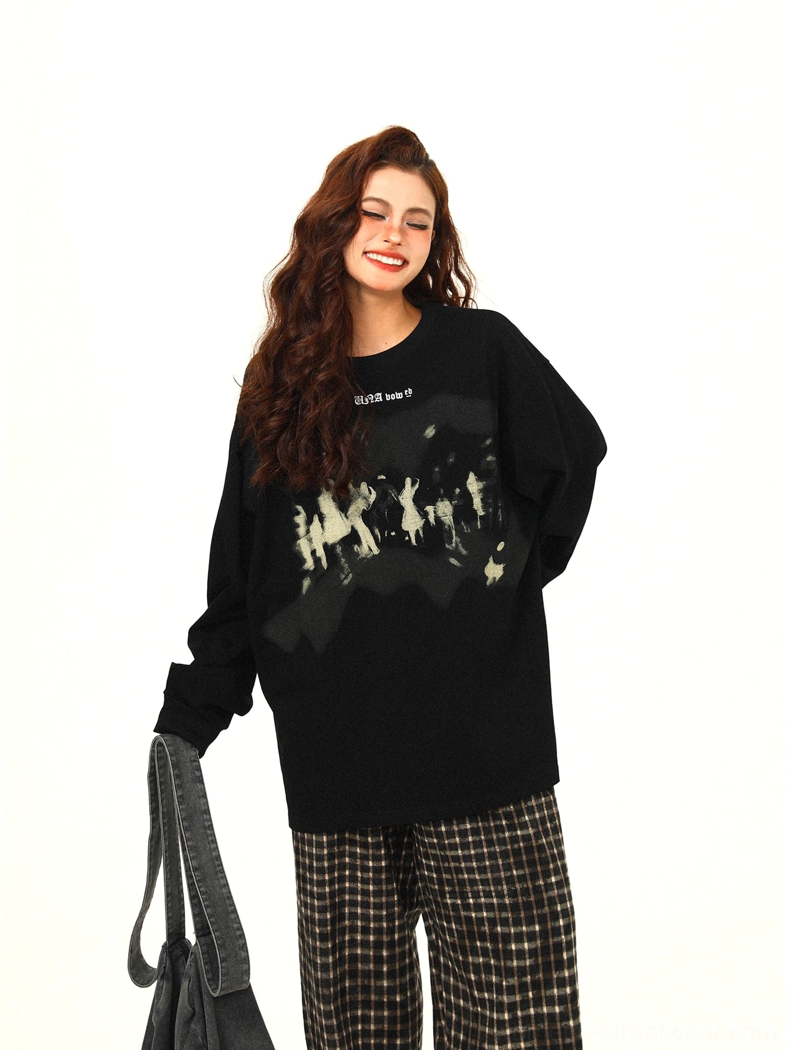 Black oversized graphic sweatshirt with small lettering above a faded crowd print, styled with checkered pants and canvas tote bag