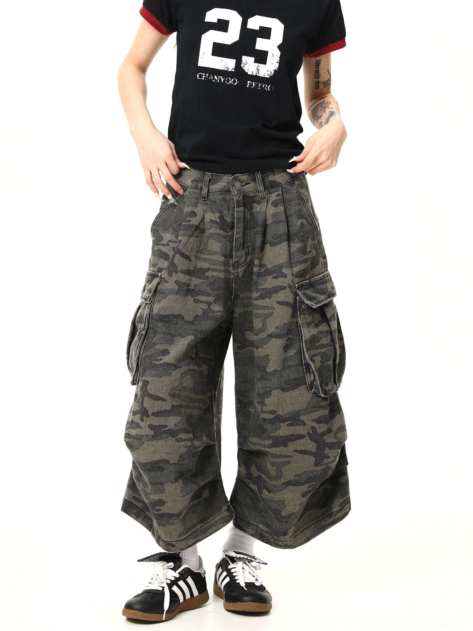 Women’s military green camo cargo pants shown from the front side with oversized cargo pockets, high-waisted pleats, cropped wide-leg shape, black graphic tee, and retro sneakers