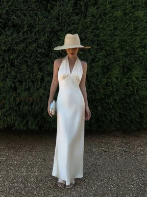 Milk apricot backless halter satin maxi dress with ruched neckline shown outdoors in a garden setting