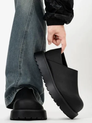 Matte black platform slip-on clogs worn with loose blue jeans and black jacket in a streetwear outfit