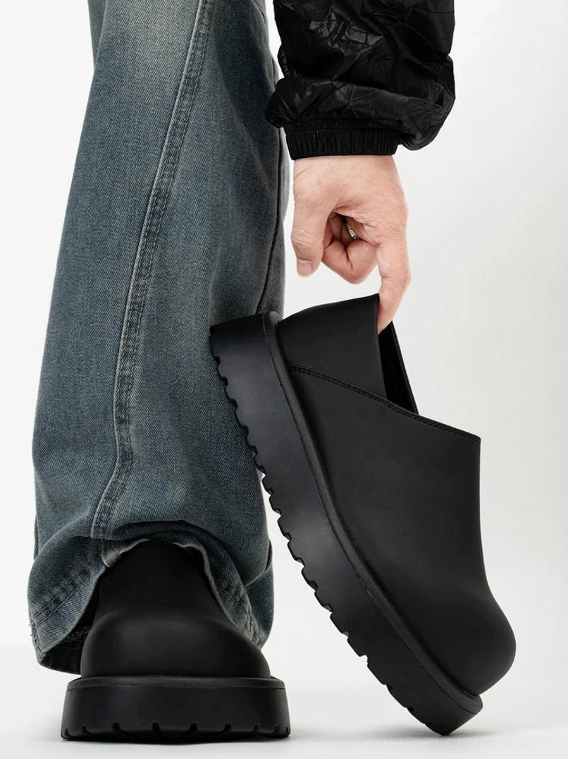 Matte black platform slip-on clogs worn with loose blue jeans and black jacket in a streetwear outfit