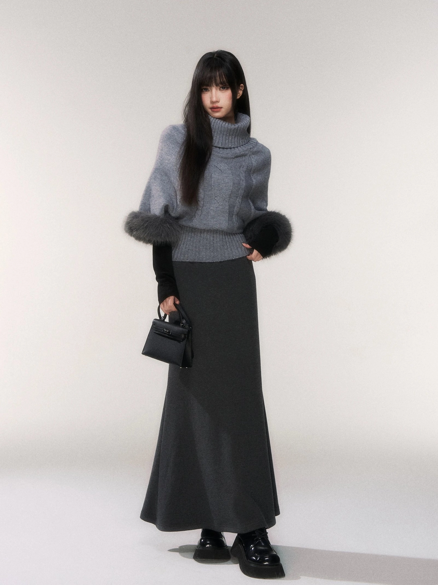 Dark gray minimalist knit fishtail maxi skirt styled with a heather gray turtleneck sweater and black handbag in a studio full body view