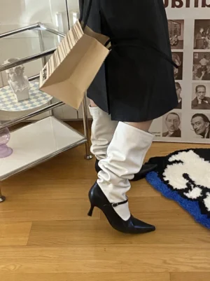 Model wearing black and white two-tone pointed-toe knee-high boots with relaxed white shaft, black pointed heel, ankle buckle strap, and pleated skirt