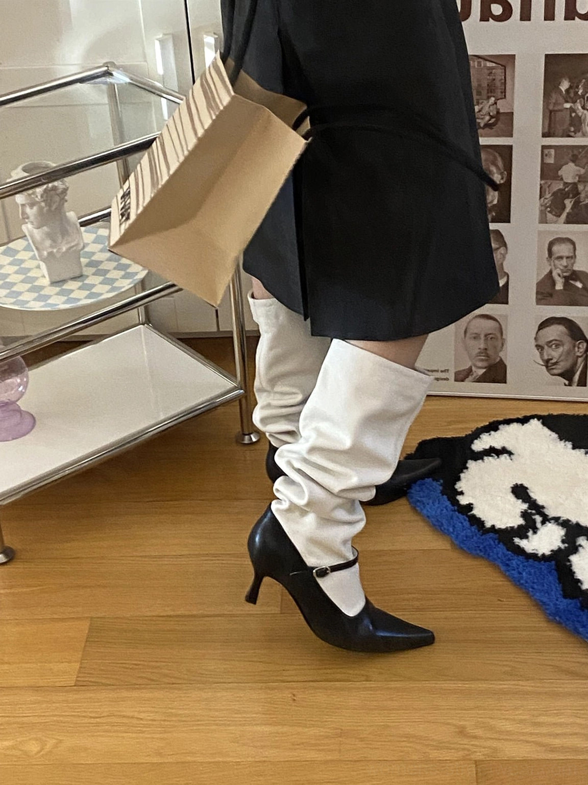 Model wearing black and white two-tone pointed-toe knee-high boots with relaxed white shaft, black pointed heel, ankle buckle strap, and pleated skirt