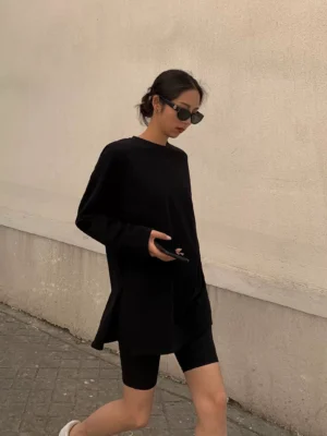 Black oversized long sleeve t-shirt dress shown from the side on a woman outdoors, featuring a loose silhouette, crew neckline, side slits, black biker shorts, and casual Seoul streetwear styling