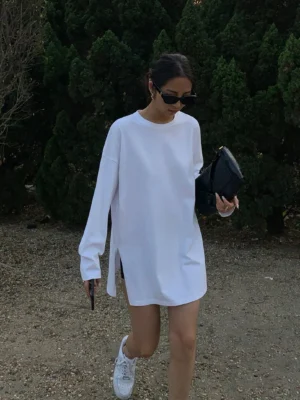 White oversized long sleeve t-shirt dress on a woman walking outdoors, featuring a crew neck, dropped shoulders, side hem slits, relaxed mid-thigh streetwear fit, white sneakers, and black clutch