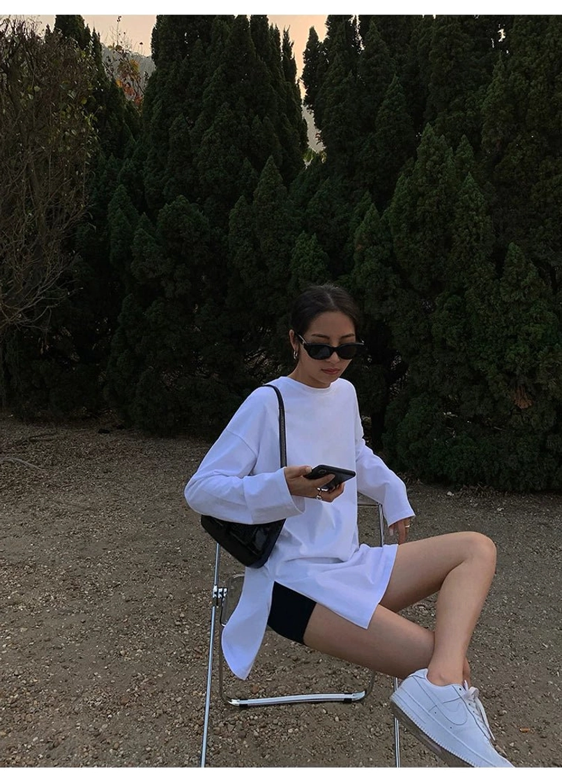 Woman sitting outdoors in a white oversized long sleeve t-shirt dress with black biker shorts, white sneakers, shoulder bag, and smartphone for an everyday Korean street style outfit