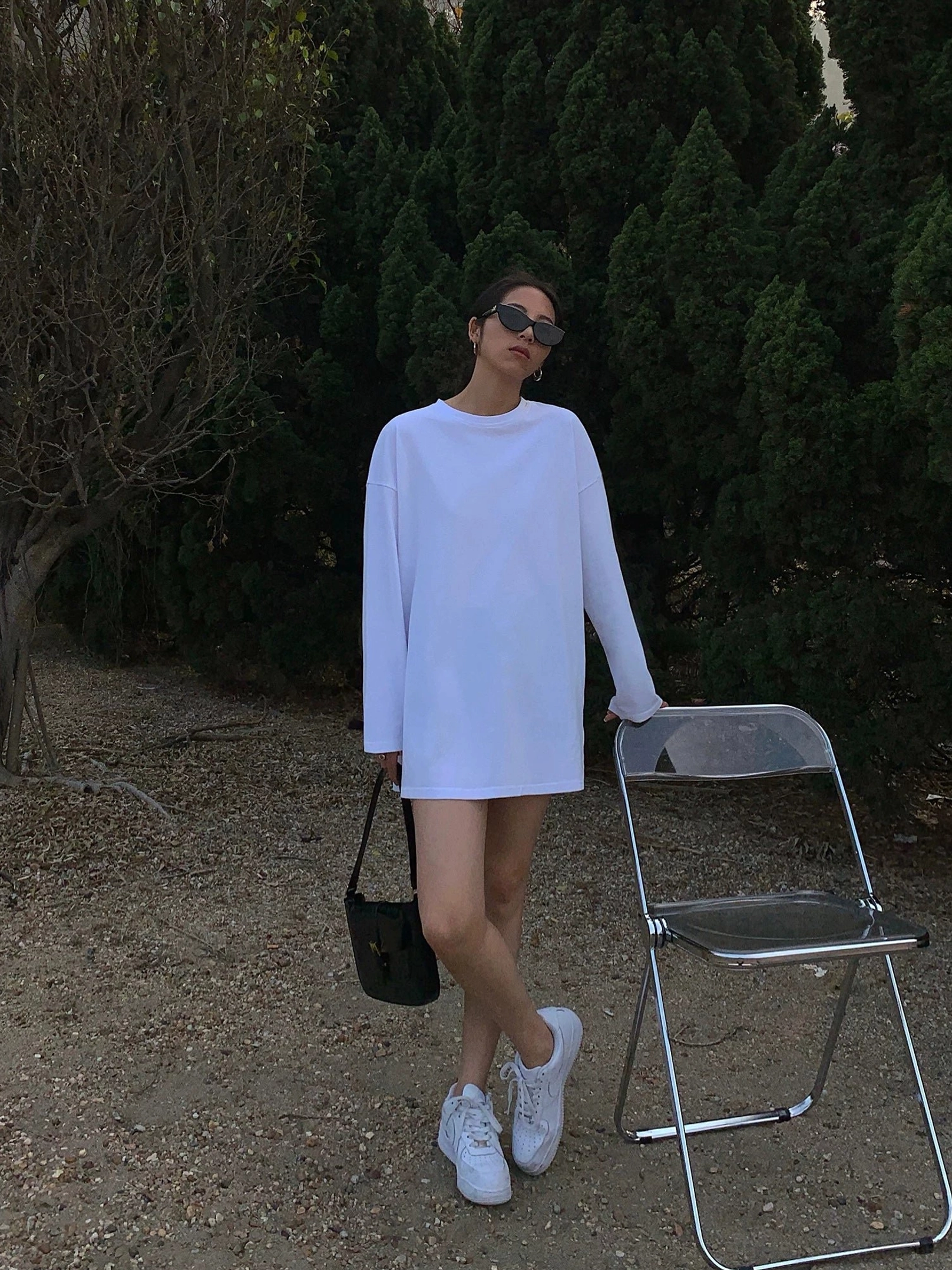 Woman wearing a white oversized long sleeve t-shirt dress in a full-body outdoor portrait, with a classic crew neck, extended sleeves, side slits, black handbag, and white sneakers