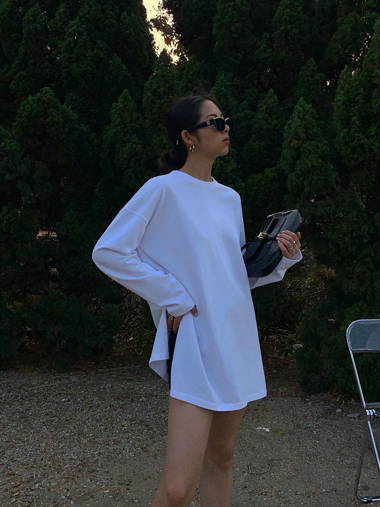 White oversized t-shirt dress with long sleeves and side slits shown in a three-quarter view, styled with white sneakers, sunglasses, and a black clutch for minimalist Korean street fashion