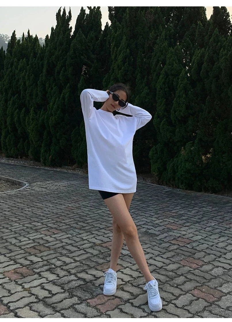White oversized long sleeve t-shirt dress styled with black biker shorts and white sneakers, showing a relaxed loose fit, side hem slits, and casual contemporary streetwear appeal