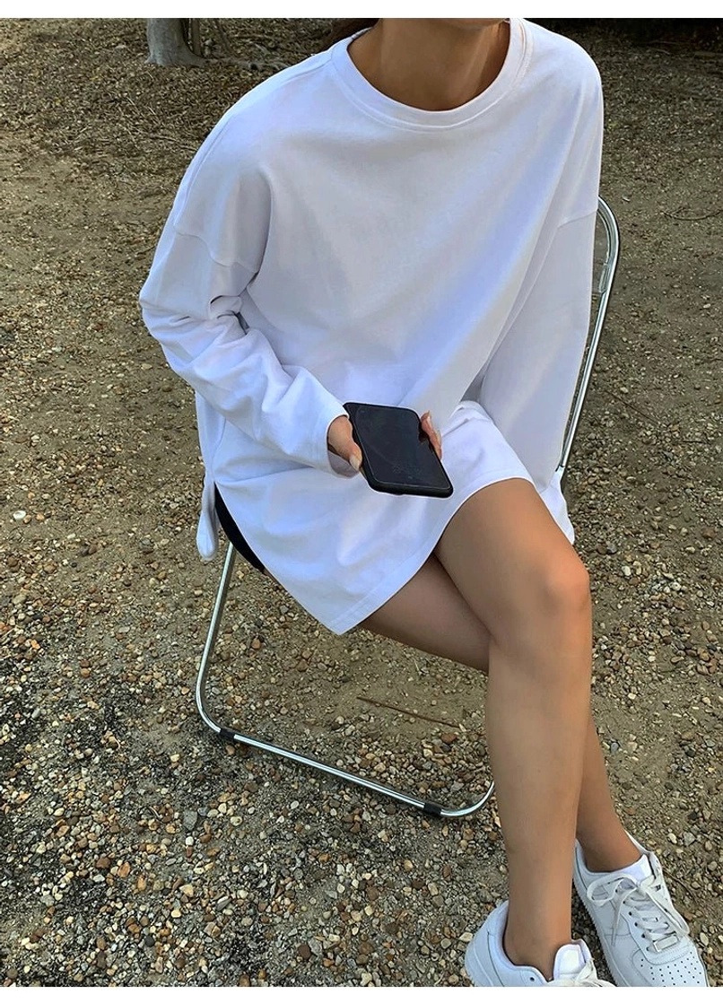 Close-up view of a white oversized long sleeve tee dress showing the crew neckline, dropped shoulder sleeve, side hem slit, loose fabric drape, and minimalist streetwear construction