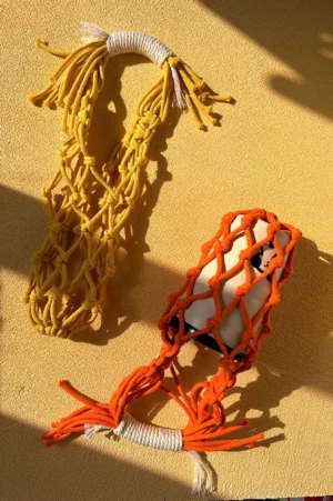 Mustard yellow and orange macrame water bottle carriers displayed with a small tumbler, showing colorful woven cotton rope and tassel handles
