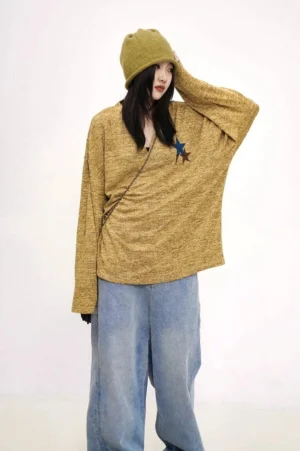 Three-quarter view of a relaxed mustard yellow oversized knit sweater with contrasting star patches, wide-leg jeans, bucket hat, and crossbody bag