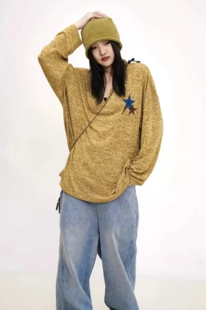 Full-body front view of an oversized mustard yellow star patch knit V-neck sweater styled with light wash jeans, a matching bucket hat, and a crossbody bag