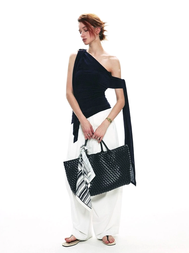 Three-quarter view of a navy asymmetric velvet top with bare shoulder, flowing side drape, white wide-leg pants, and woven tote