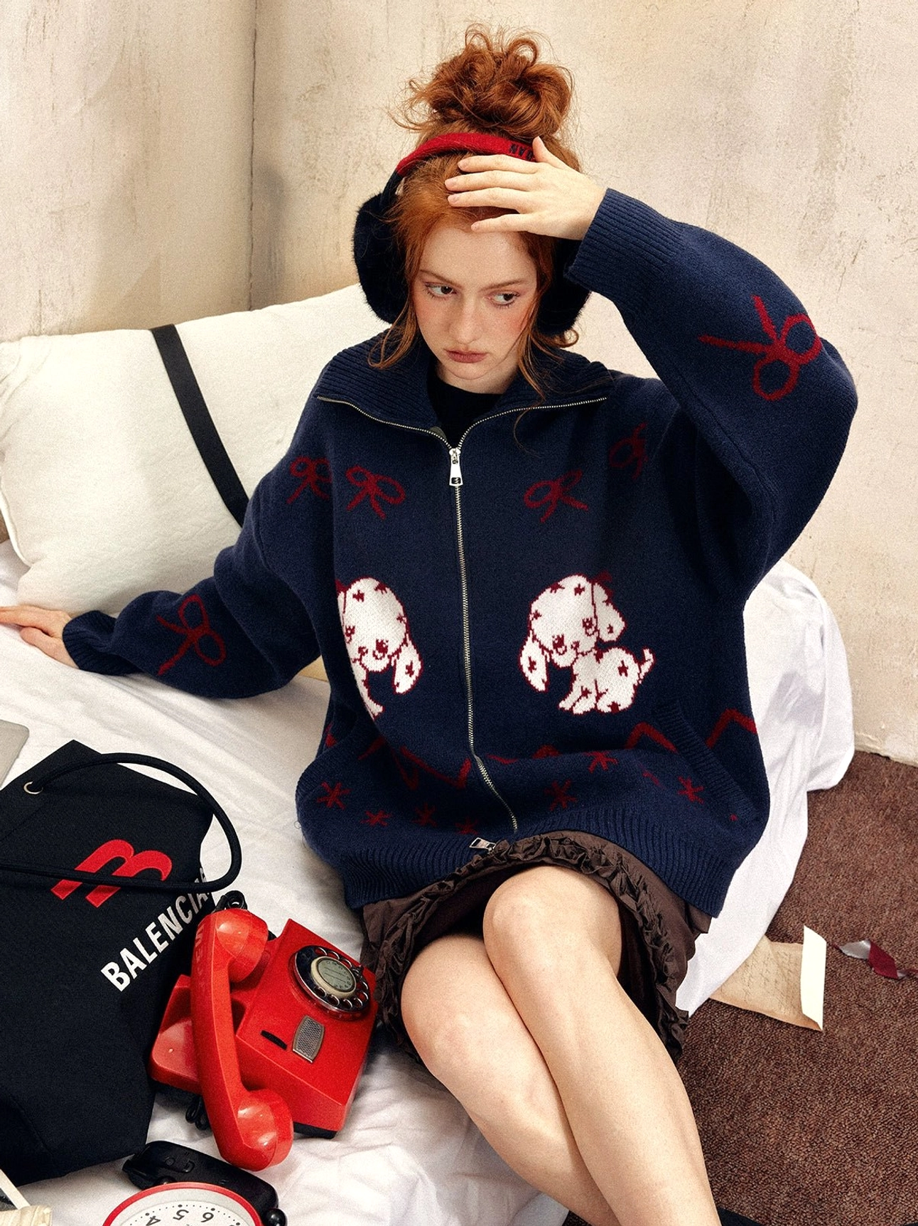 Navy blue oversized puppy print zip-up sweater with red bow and zigzag jacquard details styled with brown ruffled shorts on a vintage bed