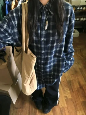 Model wearing a navy plaid oversized flannel shirt with wide-leg striped pants, feather pendant necklace, and khaki tote bag in a relaxed Korean streetwear outfit