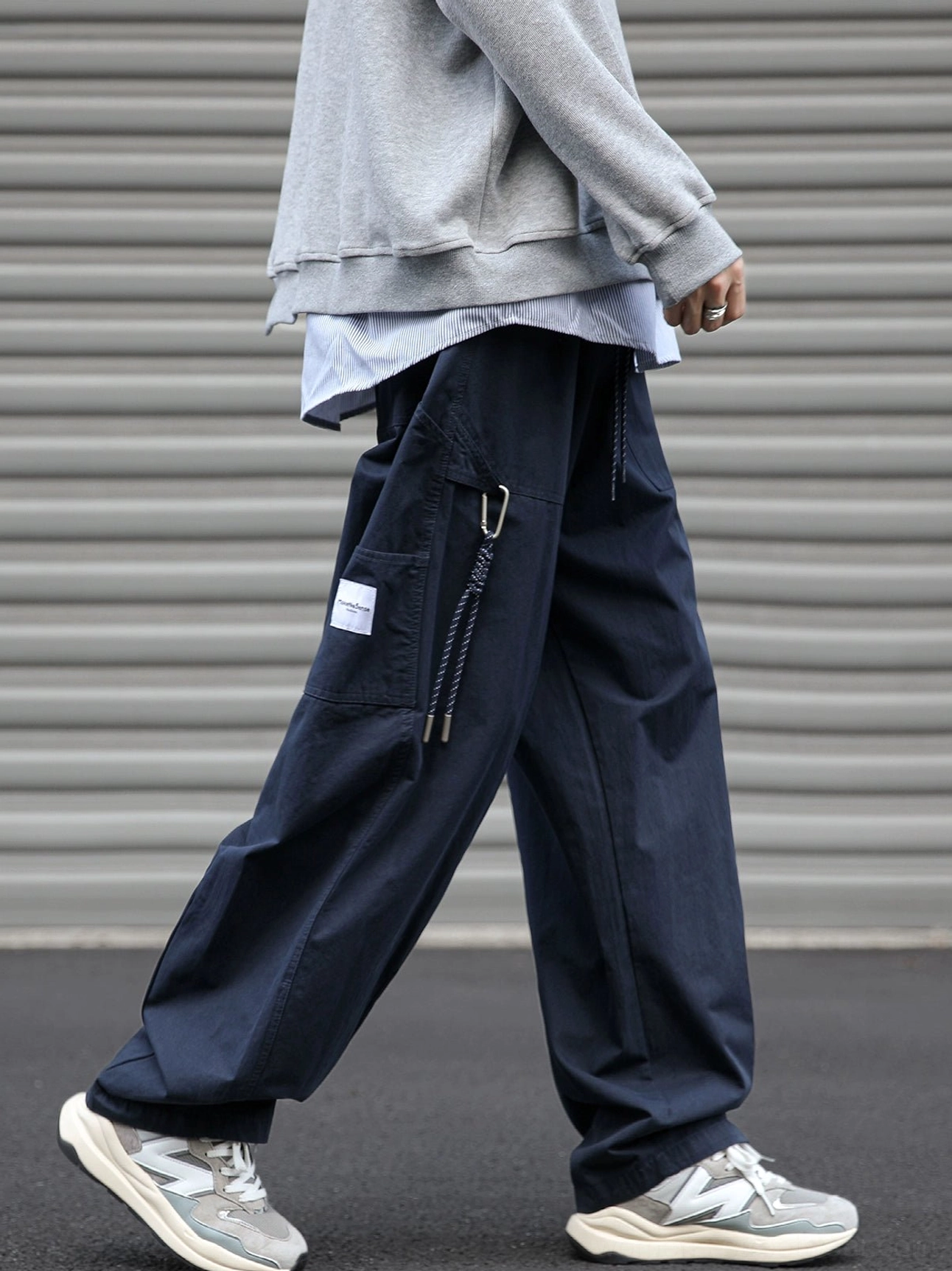 Navy relaxed utility cargo pants shown from the left side with oversized drawstring cargo pocket, wide straight-leg fit, layered gray hoodie, and retro sneakers