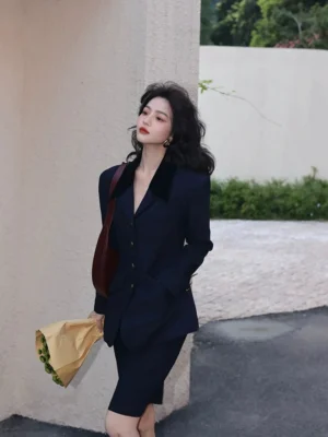 Front three-quarter view of a woman wearing a navy velvet-collar blazer and pencil skirt suit set with gold-tone buttons in a retro streetwear setting