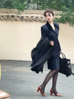 Side walking view of a woman styling a navy velvet-collar blazer and skirt set with polka-dot sheer tights red heels pearl choker and black handbag