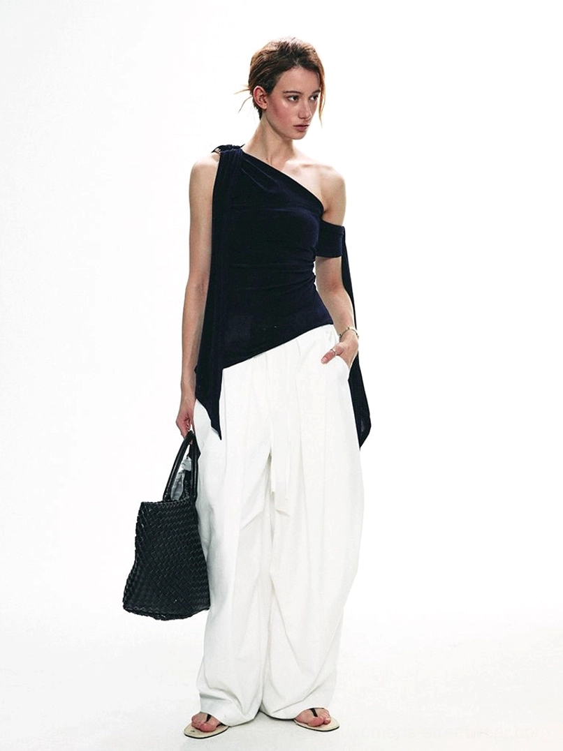 Side view of a fitted navy blue velvet one-shoulder top with asymmetric draped sash worn with white trousers and black woven tote