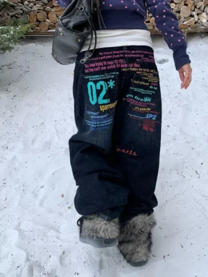 Rear low-angle view of dark indigo graffiti wide-leg jeans with colorful typography prints, styled with a cropped hoodie and fuzzy boots in a snowy streetwear setting