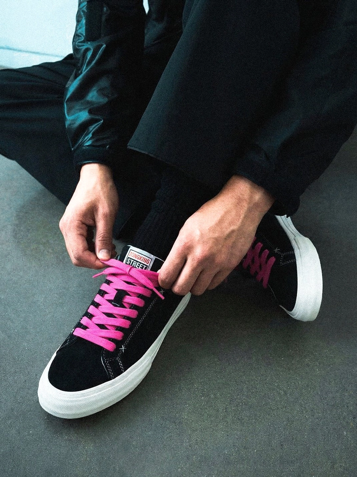 Person tying bright neon pink laces on black suede platform canvas sneakers with white rubber soles and casual streetwear styling