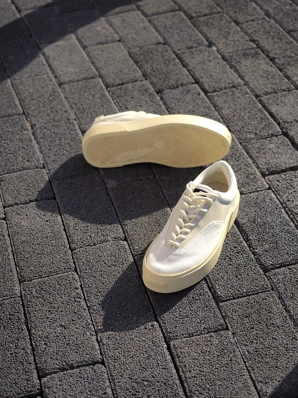 Neutral low-top platform canvas sneakers arranged on a gray brick walkway in natural sunlight, showing the off-white lace-up upper and thick creamy outsole