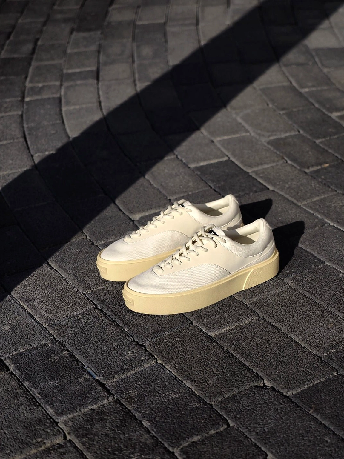 Two neutral platform canvas sneakers placed on curved gray brick pavement with dramatic sunlight shadows, styled for casual Korean streetwear