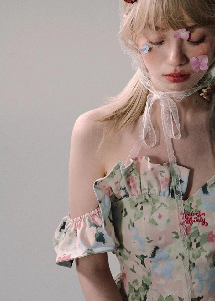 Upper-body detail of a white off-shoulder floral dress with lace-up neck ribbon, ruffled bust, ruffle sleeve edges, and decorative red chest embroidery