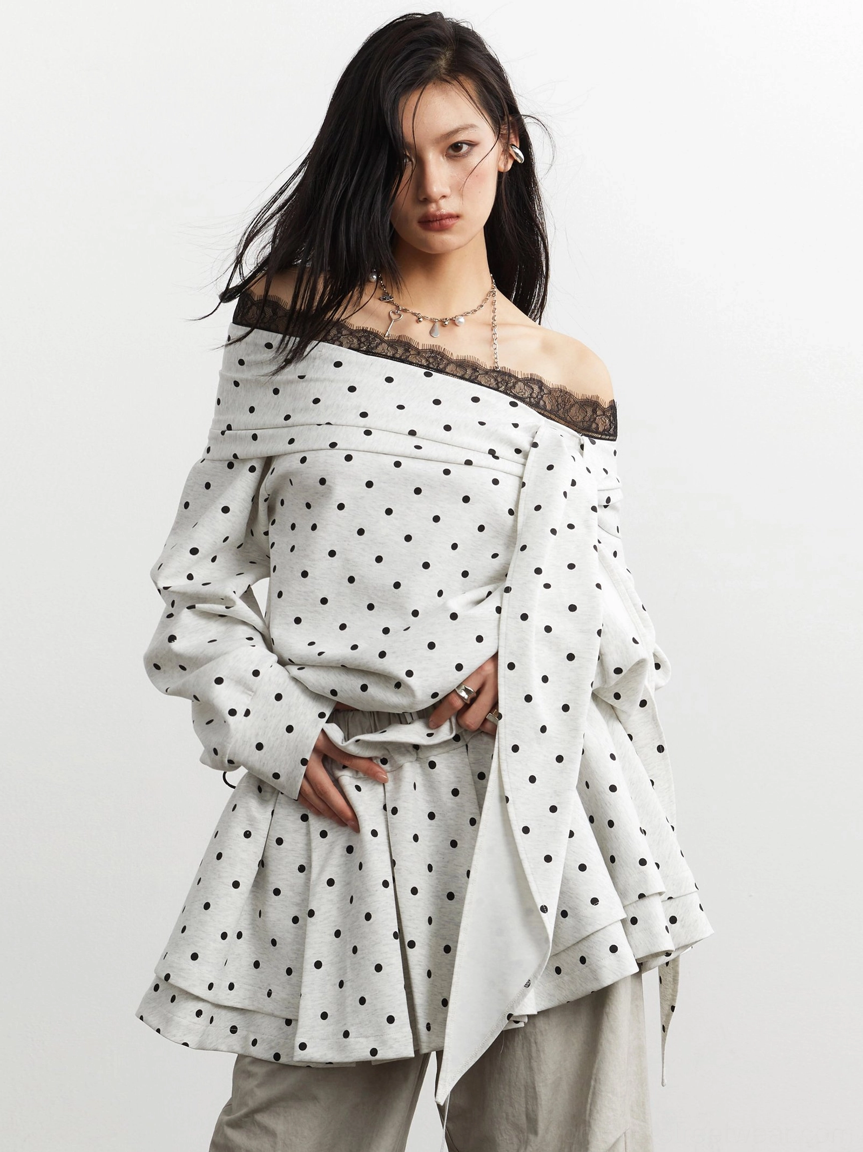 Off-white and black polka dot off-shoulder peplum top with lace trim, long sleeves, ruffled hem, and dramatic side detail styled in a minimalist studio