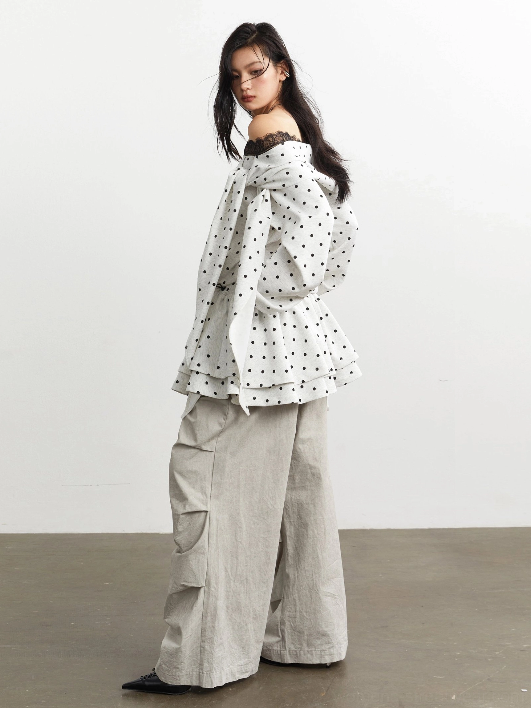 Side back view of an off-white black polka dot peplum top with large decorative bow, layered ruffled hem, long sleeves, and lace neckline