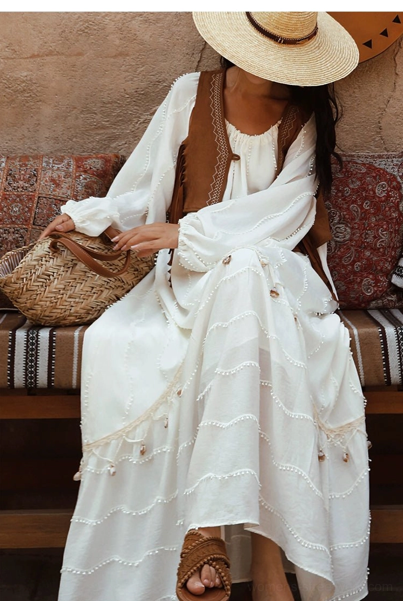 Seated close-up full-body view of an off-white bohemian maxi dress with lantern sleeves, delicate hem trim, woven straw tote, and brown sandals