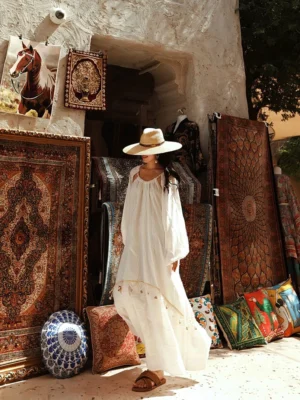 Rear three-quarter view of an off-white boho tassel maxi dress with flowing tiered skirt and voluminous lantern sleeves styled with a straw hat in a vintage textile market