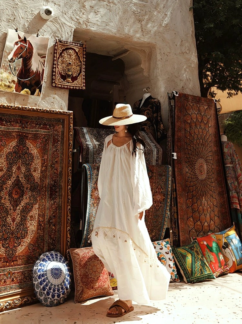 Rear three-quarter view of an off-white boho tassel maxi dress with flowing tiered skirt and voluminous lantern sleeves styled with a straw hat in a vintage textile market