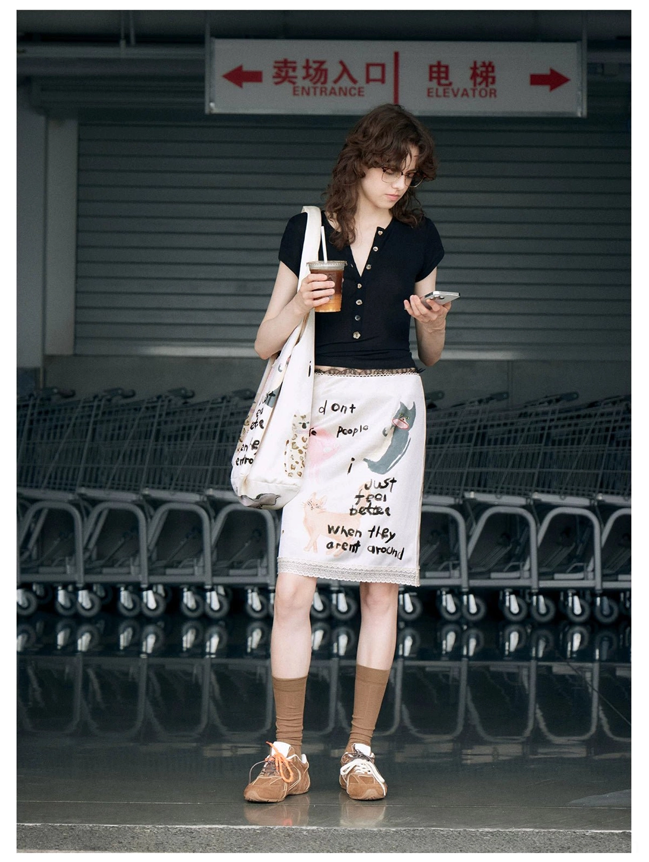 Off-white cat slogan hobo tote bag styled with a matching printed skirt while the model carries iced coffee and uses a phone
