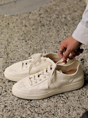 Top-down view of off-white cowhide leather sneakers with flat round laces, textured leather finish, and a hand adjusting the shoelaces