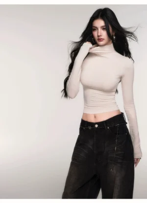 Woman lifting the collar of an off white fitted mock turtleneck crop top with fine ribbed knit texture and black wide leg jeans