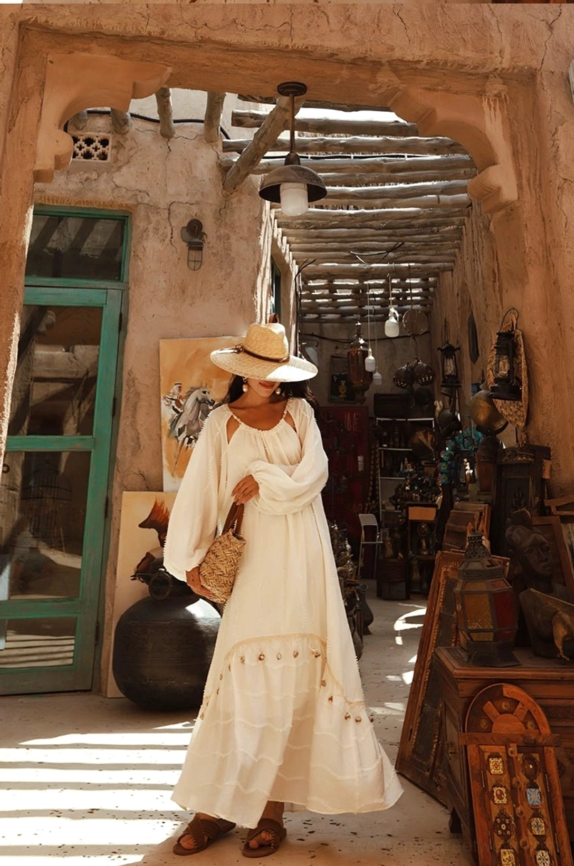 Side three-quarter view of an off-white flowing boho maxi dress with loose lantern sleeves and tiered skirt styled with brown leather sandals