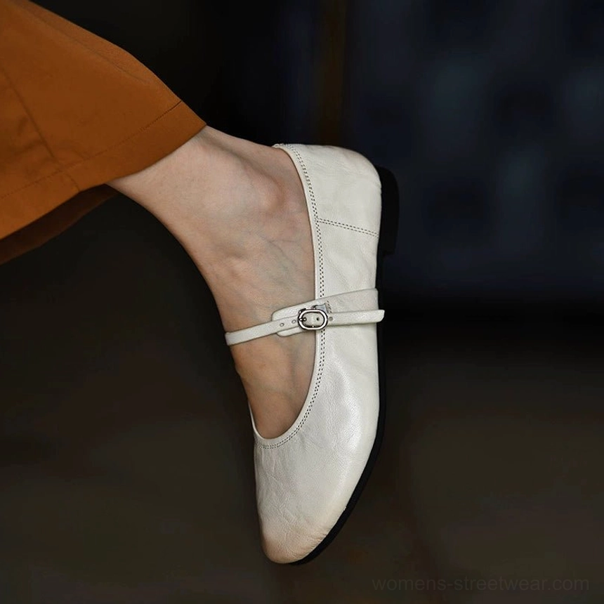 Back side view of an off white leather Mary Jane flat worn with a caramel skirt, showing the sleek upper, secure buckle strap, and low flat heel