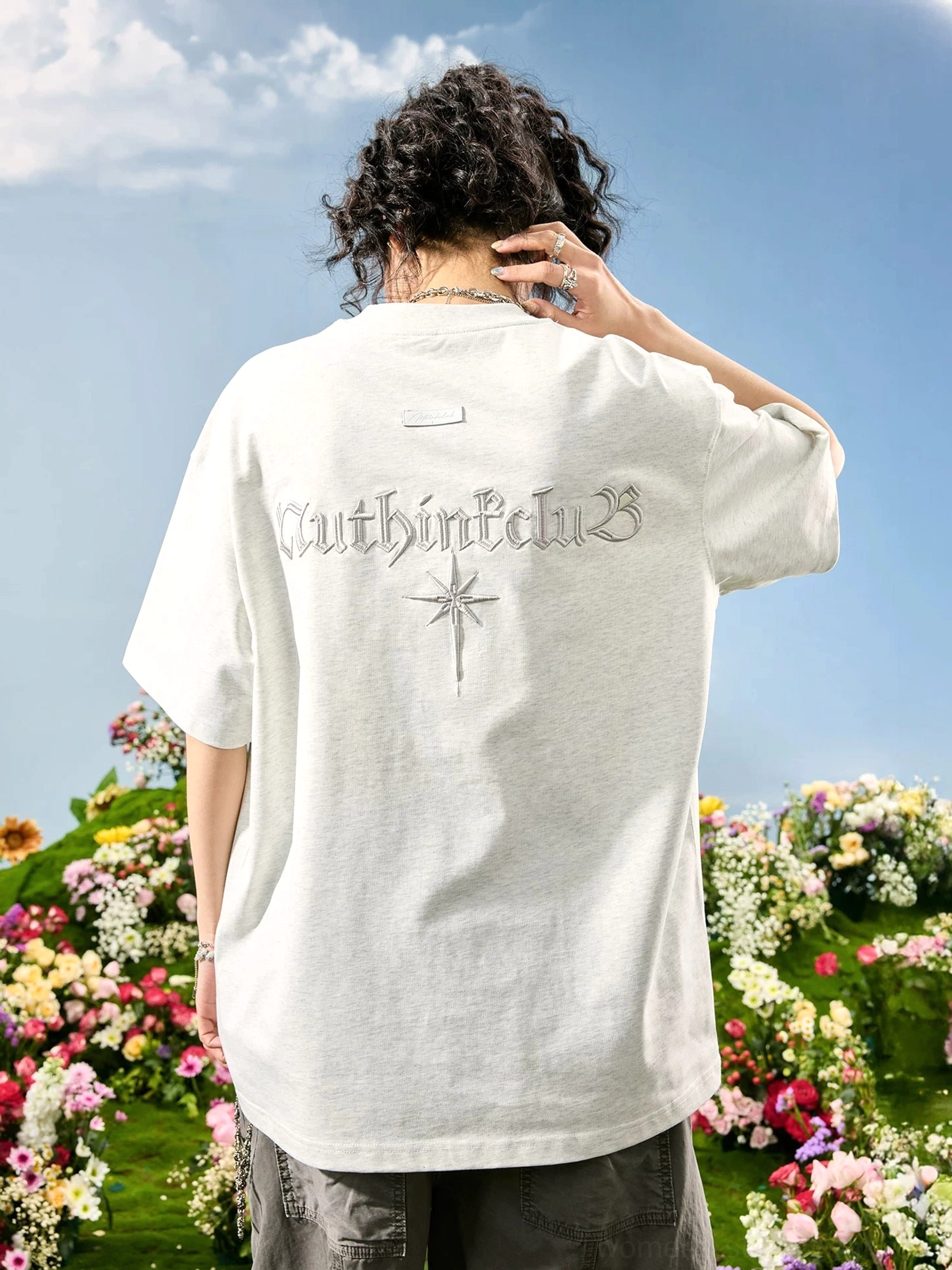 Off-white oversized Gothic embroidered back T-shirt on curly-haired female model in a flower field, showing tonal lettering and starburst streetwear motif
