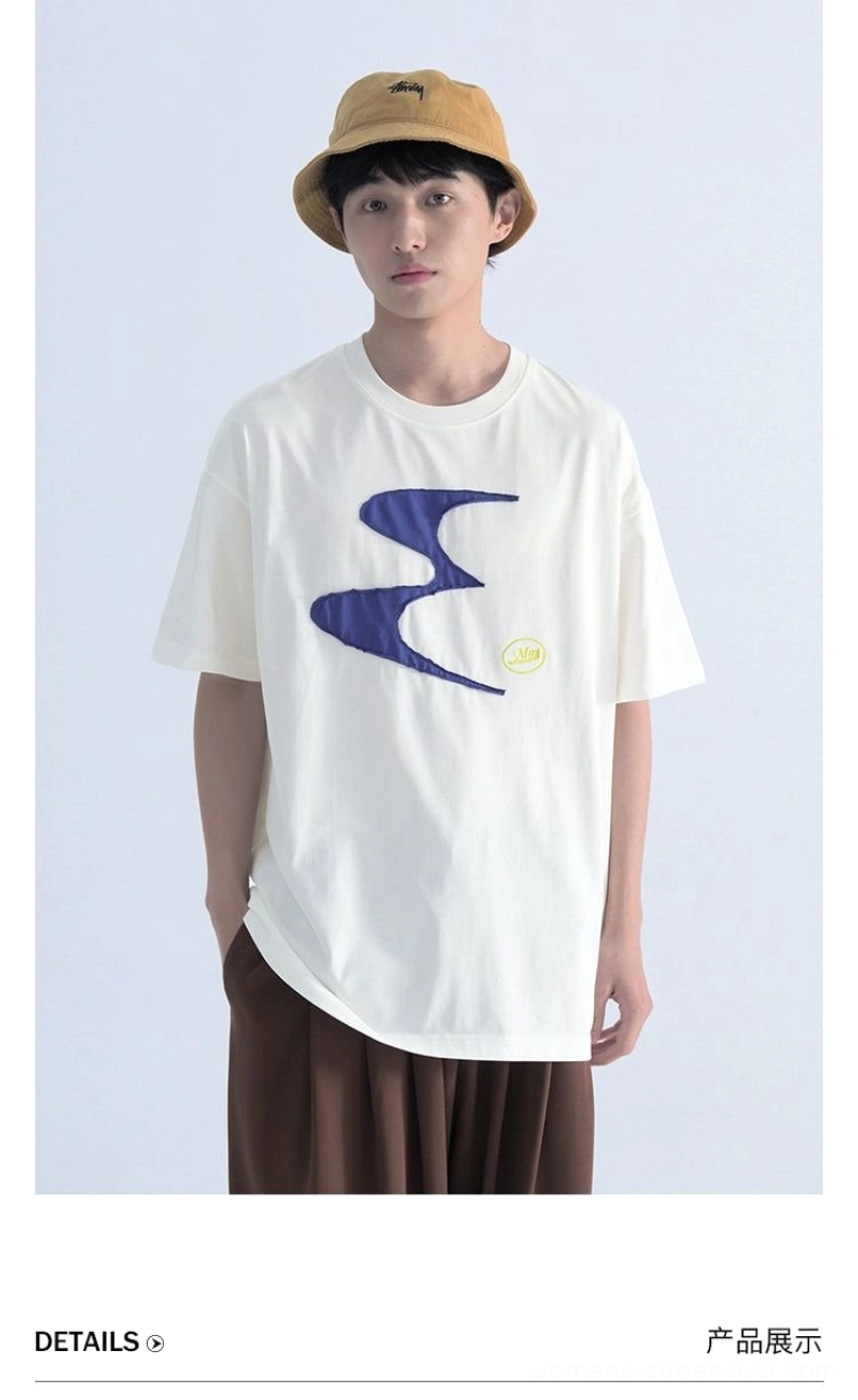 Front three-quarter view of an off-white oversized streetwear tee with navy wavy contrast graphic, styled with brown wide-leg trousers and a bucket hat