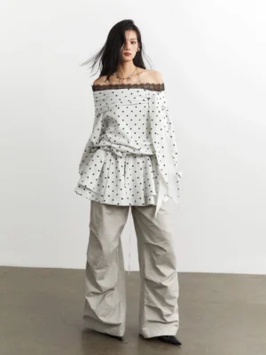 Full front view of an off-white polka dot lace-trimmed peplum top with relaxed sleeves and layered ruffles styled with beige wide-leg trousers