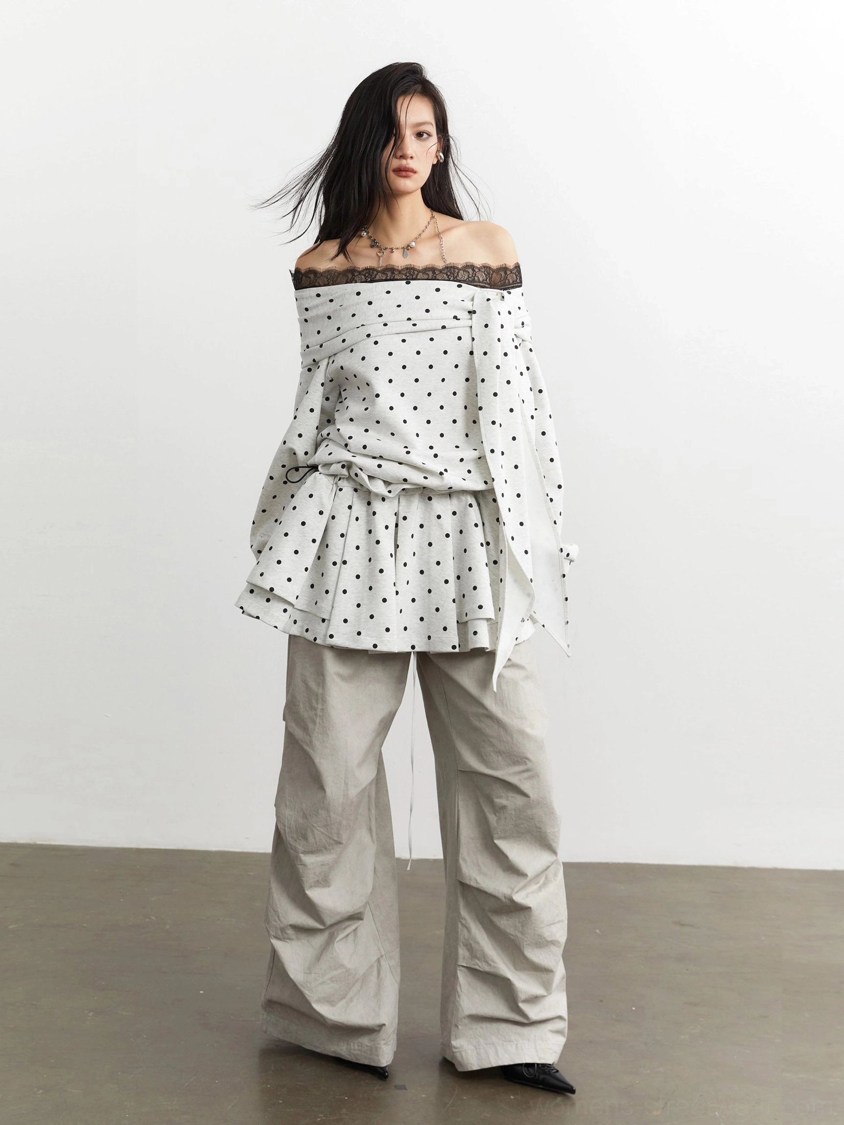 Full front view of an off-white polka dot lace-trimmed peplum top with relaxed sleeves and layered ruffles styled with beige wide-leg trousers