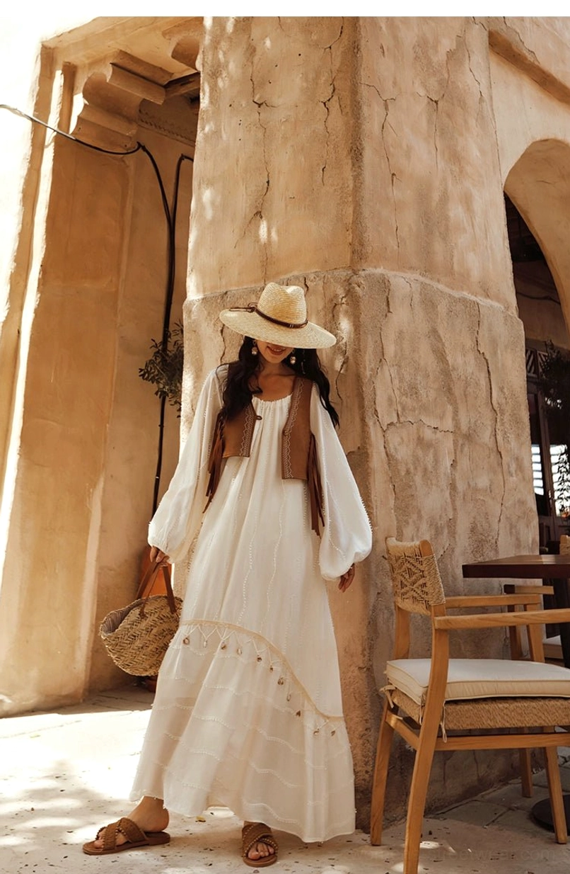 Side view of an off-white tassel maxi dress with tiered skirt and decorative hem trim layered under a brown suede fringe vest