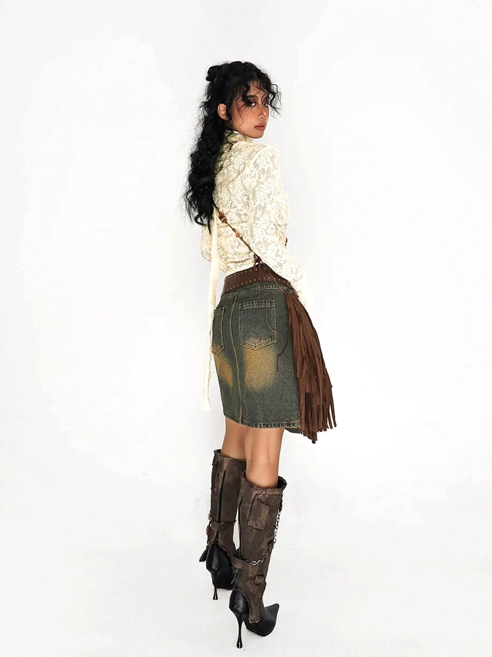 Side back view of an olive distressed denim A-line mini skirt styled with a cream lace top, fringe crossbody bag, and western brown boots