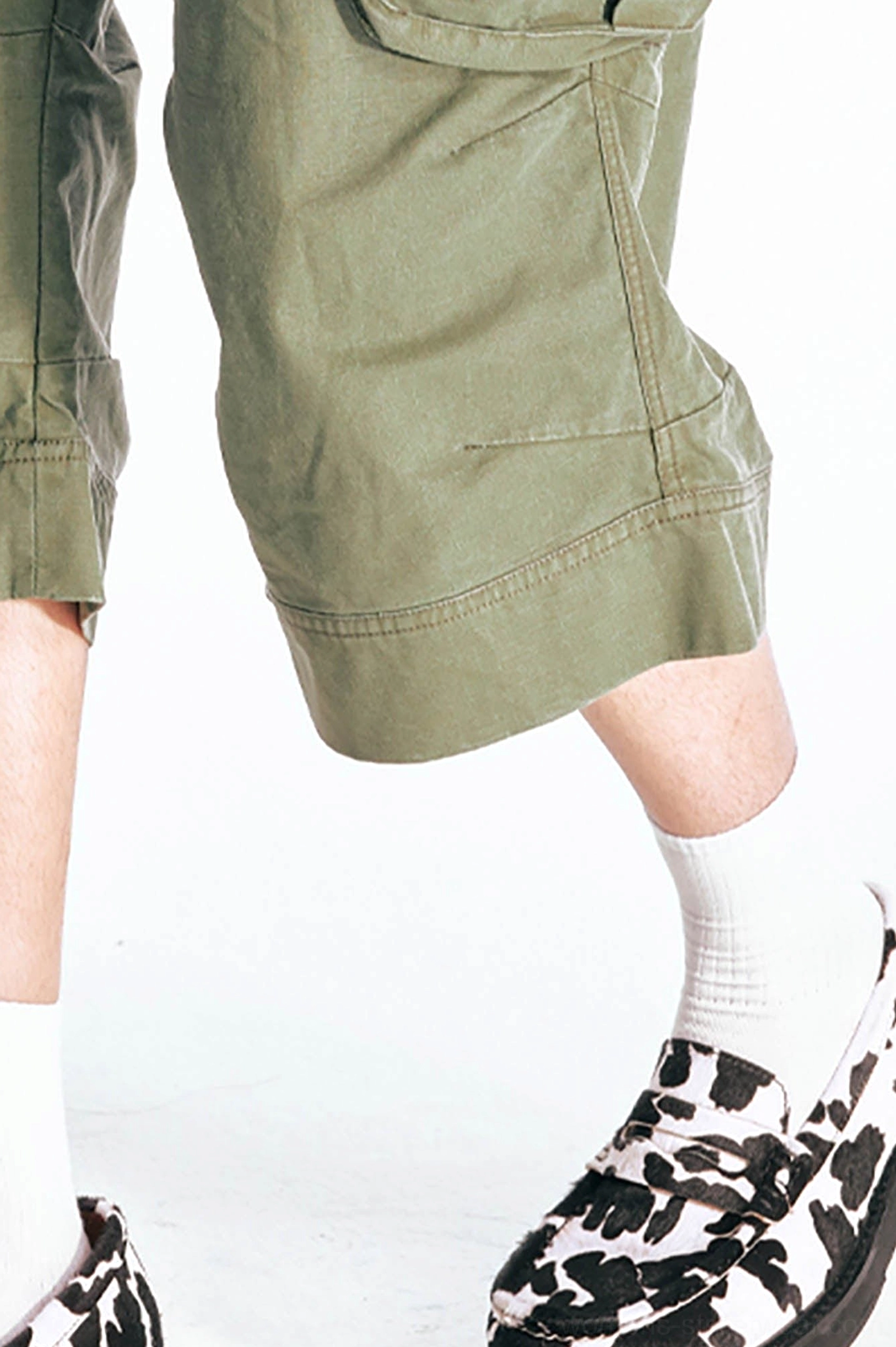 Close-up view of the wide finished hem on olive green flared Bermuda shorts with reinforced seam details, white socks, and cow-print loafers