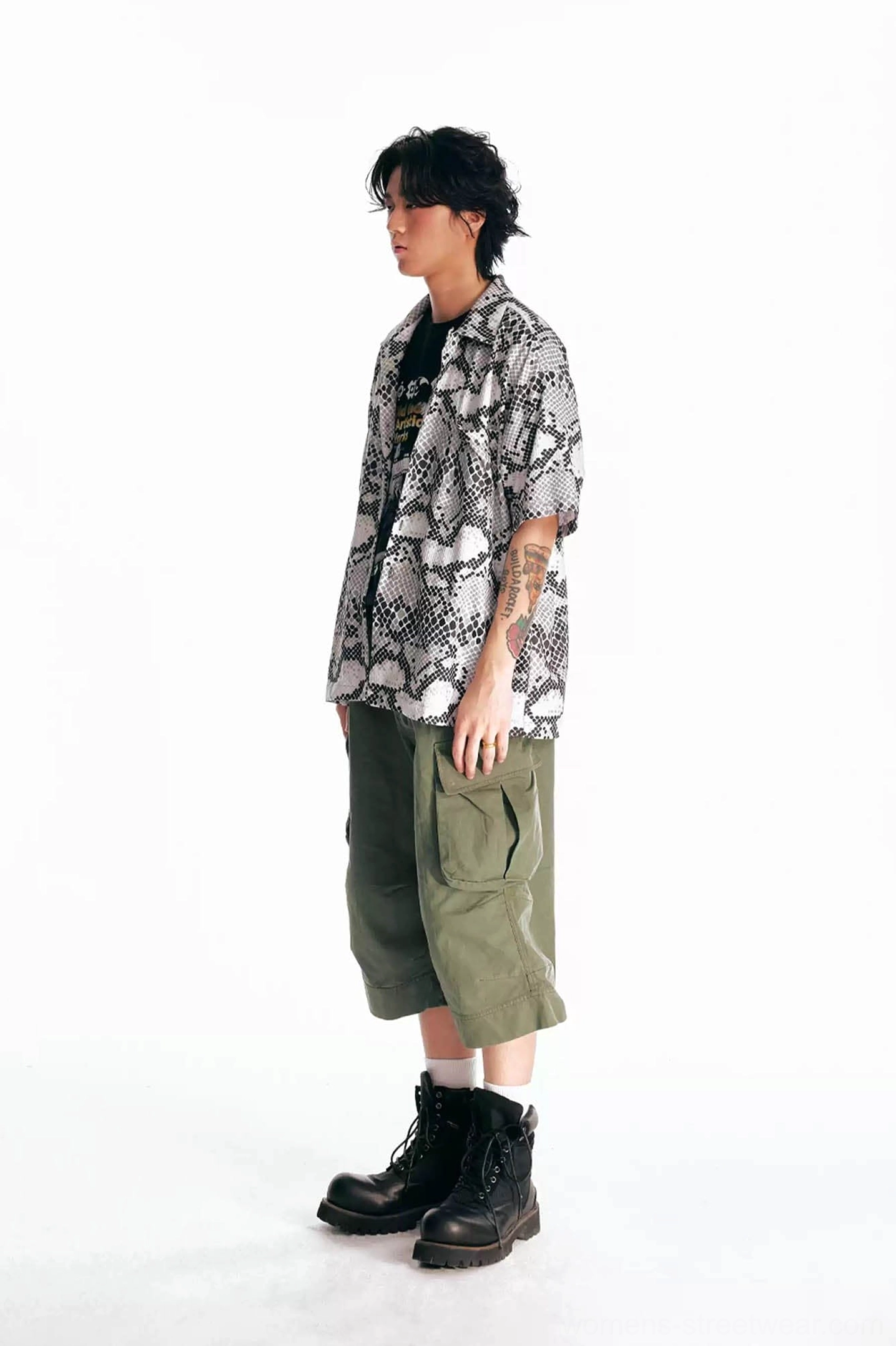 Side view of olive green flared cargo Bermuda shorts styled with a paisley shirt, black inner tee, and black work boots in a modern streetwear outfit